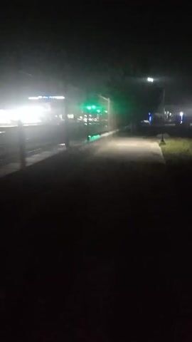 The video begins with a dark and grainy view of what appears to be a pathway leading towards a brightly lit building. There is a fence visible on the left side. As the video progresses, a person in a dark jacket stands in front of the fence. The camera then moves again, revealing a blurred building and streetlights in the background. The scene maintains a dark and unclear quality throughout.