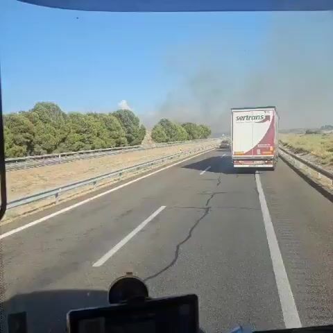 The video shows a view from inside a vehicle, likely a truck, looking out the side mirror and through the side window. The vehicle is traveling on a highway. In the foreground, the side of the truck is visible. The highway has multiple lanes and a guardrail on the side. Trees and hills are visible in the background. There are other vehicles on the highway, including several trucks and a red car. In one frame, a red truck appears to have crashed and is lying on its side off the road. There is a plume of smoke rising in the distance, possibly from the crash or a fire. The sky is clear and blue, indicating a sunny day.