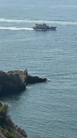 A wide shot shows a boat moving across a body of water. In the foreground, a rocky outcrop juts into the water. Several people are on the outcrop. One person is standing on the edge, and two others are beside them. Several other people are in the water near the outcrop. The water is blue and appears to be calm, with some ripples. The sky is clear and the sun is shining, creating a glare on the water. The overall scene depicts a recreational area near the sea.