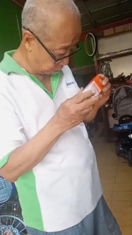 An elderly East Asian man wearing glasses, a white polo shirt with green accents, and shorts is standing in what appears to be a shop or storage area. He is holding a small orange and white box and examining it closely. The box has some text and a barcode visible. In the background, there are shelves with various items, including what looks like bicycle parts or accessories, and a blue child's bicycle or trike. The lighting suggests an indoor setting with some natural light.