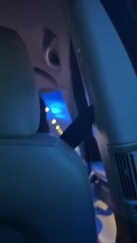 The video shows the interior of a car at night. From the perspective of the back seat, the camera focuses on the headrest of the seat in front. A pair of black cat ears are attached to the headrest. The view through the car windows shows lights passing by, indicating the car is in motion, likely on a road or highway. The interior of the car is dimly lit with a blueish hue.