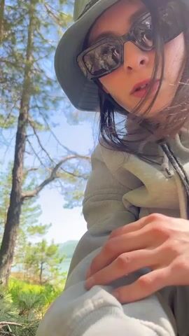 A person is featured outdoors in a scenic natural environment, likely Sliznevo, Russia. The video begins with a close-up, selfie-style shot of the person looking at the camera, wearing a green bucket hat, dark sunglasses, and a light-colored oversized hoodie. They have long dark hair. The background shows pine trees and a clear blue sky.

The camera then pans to show the person from behind as they walk a short distance to the edge of a small grassy mound. They are dressed in a casual, oversized outfit consisting of a light green hoodie, matching cargo pants, and colorful sneakers (mint green, purple, and white). The scene reveals a wide body of water, possibly a lake or river, with forested hills and mountains in the distance under a bright, sunny sky.

The person turns to face the camera, posing confidently with their hands slightly out, showcasing their full outfit. The sunlight is strong, indicating a clear, pleasant day. The setting suggests a tranquil outdoor location, possibly a forest edge overlooking a body of water, with abundant pine trees and natural foliage. There is no visible text in the video.