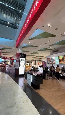 The video shows an interior view of a modern building, likely an airport terminal or large shopping area, with high ceilings and bright lighting. The floor is light-colored tile or marble. Red overhead signage with white text reads "SAO PAULO" and "DUTY FREE." On the floor, red and white markings indicate "Embarque Terminal 3 Departures" with an arrow pointing forward.

On the right side of the frame, there are several retail displays, primarily featuring cosmetics and perfumes. A tall display column features the brand "NARS" with a close-up image of a woman's face. Further down, another display has the "DIOR" logo. A few people are visible in this area, including two individuals standing near the Dior display.

In the mid-ground and background, multiple people are walking, some with luggage, indicating movement through a transit area. Large digital screens displaying colorful advertisements or images are mounted on walls in the distance. The overall scene suggests a vibrant commercial space.
