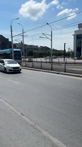 The video shows a street scene on a sunny day with a clear blue sky and some white clouds. In the foreground, a gray asphalt road is visible with a white painted line running down the center. On the right side of the road, a sidewalk and a building are partially visible. In the middle ground, a white sedan is driving on the road. Behind the car, a blue and white tram is traveling on a separate track. There are power lines overhead for the tram. Further in the background, more buildings and trees are visible. The overall scene depicts urban transportation and city life.
