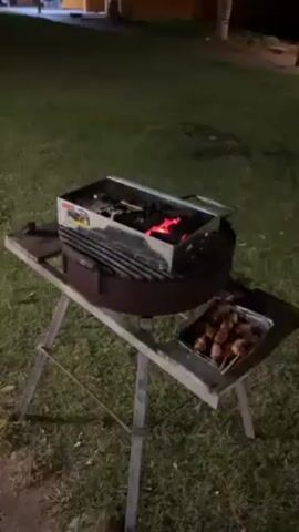 Here is a matter-of-fact description of the scene: 

The video shows an outdoor grill on a grassy lawn at night. The grill is old and rusty, resting on a wooden table. A metal container sits on top of the grill, holding glowing embers. To the side of the grill, there's an aluminum tray filled with skewers of cooked meat. The background is dark, with a building visible in the distance. There's a slight breeze, evident from the rustling sound. No people are immediately visible in the frame.