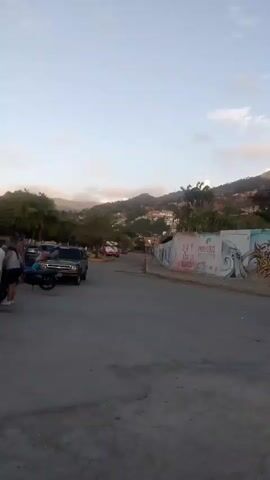 A video captures a street scene with a group of people gathered near a motorcycle and a pickup truck. The background reveals a hillside dotted with buildings and trees, under a sky with scattered clouds. The road appears paved, and a graffiti-covered wall runs along the side of the street. The overall atmosphere suggests an outdoor urban or semi-urban environment, possibly during daylight hours.