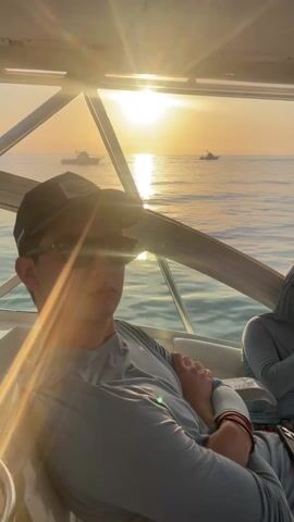 The video shows people on a boat, likely fishing. The sun is setting or rising, creating a warm glow.  Initially, two men are visible in the boat's cabin.  The camera pans to show other people setting up fishing gear on the back of the boat. Fishing rods are prominently displayed. The boat is on the ocean, with land visible in the background.  Other boats are in the distance.