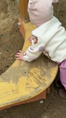A baby wearing a pink hat, cream-colored sweater with flower-shaped designs, and purple pants and shoes is shown. The baby is positioned near a yellow, curved structure, and rests their hands on the surface, which is covered with dirt. The background consists of soil and the base of the yellow structure, partially supported by bricks.