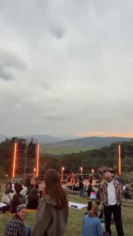 The video shows an outdoor music festival during twilight. A crowd of people are gathered on a grassy hillside, facing a stage with a band performing. Two large speaker towers with bright orange lights flank the stage. The sky is cloudy with hints of orange and pink from the setting sun. The landscape behind the stage consists of rolling hills covered in trees. Some people are sitting on blankets, while others are standing or walking around. There are also some tents set up in the background. The overall atmosphere appears relaxed and festive.