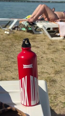 A red aluminum water bottle with a black lid sits on a white surface in the foreground. The bottle has a white sticker with text and a silhouette of buildings. In the background, a body of water is visible under a blue sky with scattered clouds. Two empty white lounge chairs are positioned on a sandy and grassy area near the water. One lounge chair has towels and other items on it. The legs and torso of a person are visible, reclining on one of the lounge chairs. Another person is partially visible further in the background. The environment appears to be a beach or lakeside setting on a sunny day.