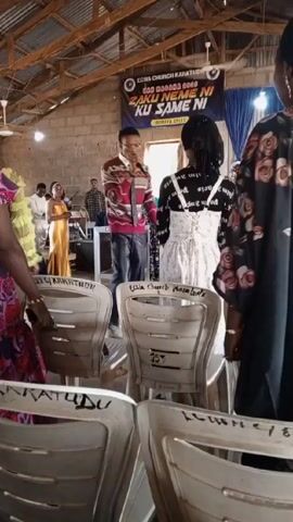 The video shows a church service taking place indoors. A banner reading "ECWA CHURCH KARATUDU DAN MAGANA 2023 ZAKU NEME NI KU SAME NI JERIMIYA 29:13" hangs in the background. A man in a red sweater stands at a podium speaking into a microphone. To his left, women are standing behind a table with a blue container on it. In the foreground, several white plastic chairs are visible. The church has brick walls and a wooden beam roof. There are several people present, including musicians with instruments. The atmosphere appears to be one of worship and communal gathering.
