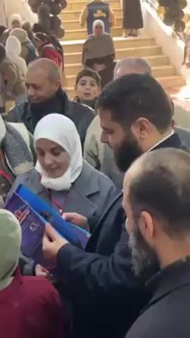 The video captures a crowded indoor scene. A woman wearing a white hijab and grey coat is holding a blue book open, showing it to two men standing close to her, both of whom have beards. Other people are crowded around, including a man in a black jacket who is smiling at the camera. In the background, there's a staircase, with people walking up it. There are also some balloons visible. The atmosphere appears busy and engaging, with several individuals focused on the book.