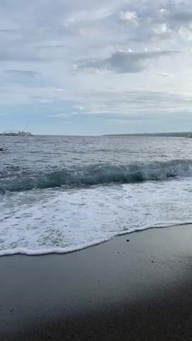 A video shows a beach with dark sand and gentle waves rolling onto the shore under a cloudy sky. In the distance, a boat is visible on the horizon. The video is shot from a low angle, close to the sand, and focuses on the movement of the water and the texture of the beach. The overall mood is calm and serene.