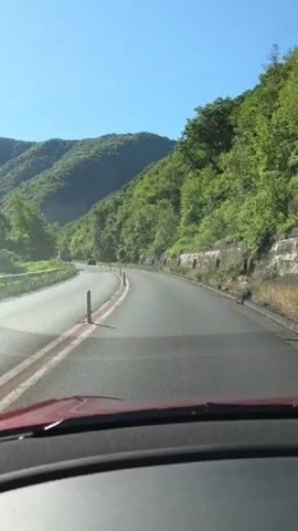 The video is a first-person perspective of driving on a paved road that curves to the left through a mountainous, forested area. The road has two lanes with red and white lane markings. Orange traffic cones are visible along the road. The sky is clear and blue. The surrounding mountains are covered in lush green trees. The dashboard of a red vehicle is visible in the foreground. There are a few cars in the distance on the road ahead. The lighting suggests it is daytime, likely morning or afternoon.