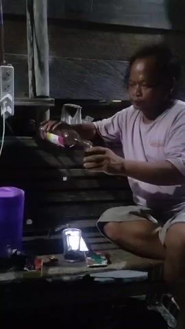 A person is sitting on a wooden surface, wearing a pink t-shirt and shorts. They are holding a glass bottle with a pink liquid in their right hand and pouring it into a clear glass. There is a small lamp with a light bulb next to the glass. In the background, there is a wooden wall and some electrical outlets. The scene is dimly lit.