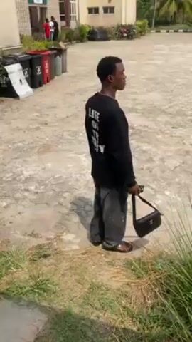 The video shows a young man standing on cracked pavement, looking into the distance. He is wearing a black long-sleeve shirt with writing on the back, gray pants, and sandals. He is also carrying a small black handbag. He puts his hand up to his forehead as if to shield his eyes from the sun, and then raises his arm to point something out. In the background, there are trash cans, a building, and two other people standing near the building. There is also a motorcycle in the distance. The surrounding area appears to be outdoors, with some greenery visible.