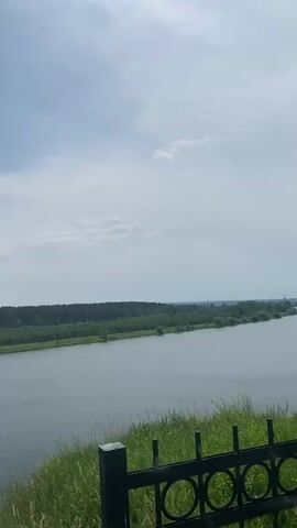 A still, wide shot of a river on a partly cloudy day. The river fills the middle of the frame, with green foliage and a dark metal fence in the foreground. A dense line of trees runs along the far bank of the river, with a few distant structures visible on the horizon. The sky is a pale blue with scattered white clouds.