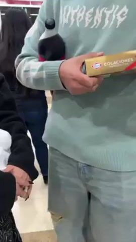 A group of people are standing in a convenience store, possibly waiting to pay at a checkout line. One person is holding a white plastic bag filled with items, and another is holding a box of "COLACIONES" brand wafer cookies. There is a wooden shelf with eggs and other food items. The overall lighting is bright.