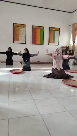 A group of four young females, dressed in Islamic attire including headscarves, are performing a coordinated dance. They are on their knees in a room with tiled flooring. In front of each dancer is a circular, shallow basket. The walls are white, and three large framed charts with text and colored blocks are visible. One dancer in the front is wearing a pink top and brown skirt, while the other three are in dark-colored outfits. The dancers' arms are extended outwards and slightly upwards.