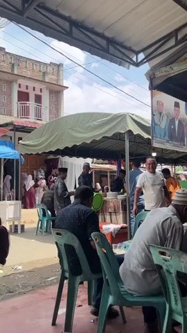 The video shows an outdoor gathering with several people sitting at tables under a canopy. In the foreground, two men are seated at a teal table with plastic chairs. The man on the left is wearing a white shirt and a white skullcap, and the man on the right is wearing a black shirt. There are plastic cups and bottles on the table. In the background, more people are seen standing and sitting, some under another green tent. There are food serving stations and decorations visible. A banner with portraits of men and text in a foreign language hangs from the top. The environment appears to be a street market or an outdoor event.