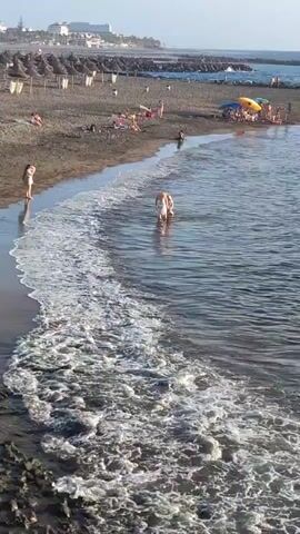 A vertical, high-angle, eye-level, medium shot shows a crowded beach with black sand on a sunny day. In the foreground, the ocean water laps against dark, rocky shoreline. In the middle ground, two people wading in the shallow water are visible. The person in front is wearing a white swimsuit and the person behind them is wearing a light-colored swimsuit. Both are facing away from the camera. In the background, a sandy beach is lined with rows of thatched umbrellas and lounge chairs. Several people are visible sunbathing and walking along the shoreline. A few more people are further up the beach near buildings. The sky is clear and light blue.