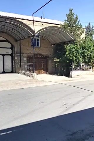A video shows a residential building with a large arched carport covering a driveway. The building is made of brick and has a white roof. There are trees and greenery around the building. The video is shot from a street in front of the building. The time stamps indicate the video lasts only a few seconds.