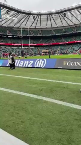 The video shows a low-angle view of a rugby field within a large stadium. The field is green and lined with white. There are spectators in the background, occupying the stadium seating. Along the edge of the field, advertising displays for Allianz and Guinness are visible. A photographer wearing a black vest and a worker in black clothing are kneeling near the advertising. Additional people, also in black, are walking further down the field. There are black cases on the sideline. The weather appears overcast. A scoreboard is visible in the upper left corner of the frame, displaying scores for England. A person wearing a dark jacket and black pants walks across the foreground, partially obstructing the view.