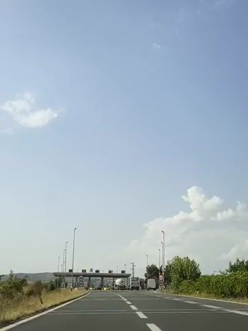 A first-person perspective video shows a car approaching a toll booth plaza on a highway. The sky is blue with scattered clouds. There are speed limit signs on the road indicating 40. Several vehicles, including cars and trucks, are visible in front of the toll booths, with some in motion and others stopped. Trees and vegetation line the sides of the road.