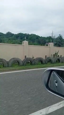 The video shows a cow grazing near a wall lined with tires. A person walks past a utility pole, and then another person walks on the side of the road near some benches and a small building. The scene is outdoors with trees and power lines in the background, under a cloudy sky. The footage appears to be taken from a moving vehicle, as indicated by the side mirror in the foreground.
