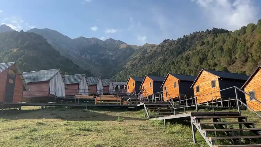 The video shows a row of wooden cabins, some painted red and white, others dark brown, nestled in a mountainous landscape. The cabins are elevated on stilts and accessed by metal staircases. The surrounding terrain is grassy with scattered trees on the slopes of the mountains, which are rocky and have patches of green vegetation. The sky is blue with some clouds. There are no people visibly present in the shot. The camera is static, offering a wide view of the cabins and the natural environment.