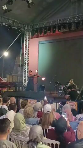A fire performer is on a stage in front of a crowd. The performer is wearing a black outfit and is juggling flaming objects. There is a drum set and other musical instruments on the stage. The crowd is seated and many people are holding up their phones to record the performance. The stage is lit by spotlights and the overall atmosphere appears to be a nighttime outdoor event.