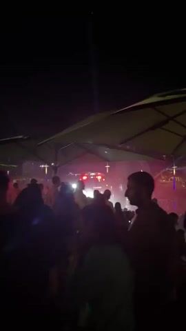The video shows an outdoor gathering at night, likely a concert or festival. Several people are visible, with some in the foreground out of focus. In the background, there is a stage with lights and a crowd of people. Large green umbrella-like structures are also present, possibly serving as shelters or decorative elements. The lighting shifts between green, red, and white, creating a dynamic atmosphere. People appear to be standing and mingling, with some possibly dancing. The overall environment suggests a lively social event taking place after dark.