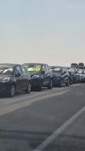 The video shows a traffic jam on a road with multiple lanes. Cars are bumper to bumper, with some people standing outside their vehicles. The sky is clear with a few clouds. The scene appears to be during the daytime, possibly in the late afternoon or early evening given the lighting. The vehicles are of various makes and models, including sedans, SUVs, and possibly a van. The road surface is asphalt and appears to be in good condition. There are lane markings visible on the road. The overall impression is one of significant congestion and delay.
