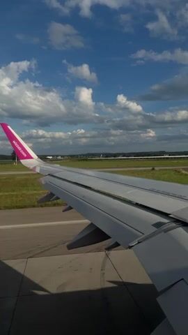 The video captures a view from an airplane window, looking at the wing and the runway outside. The wing, with its distinctive pink tip featuring the Wizz Air logo, takes up a significant portion of the frame. The plane appears to be on the ground, possibly taxiing or preparing for takeoff, as the runway is visible in the background. The sky is bright blue with scattered white clouds, indicating fair weather. The surrounding environment includes green fields and some distant buildings or structures. The lighting suggests it is daytime, likely morning or afternoon. There are no people visible in the frame. The camera perspective is static, suggesting it was filmed from a stationary position inside the aircraft.