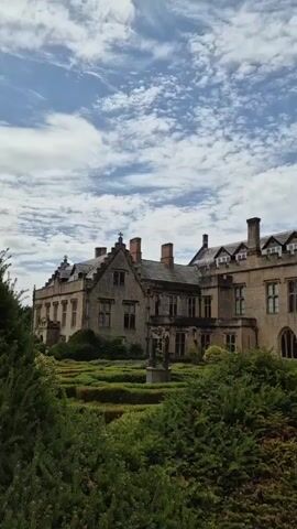 The video shows a drone footage of a historic stone building with a manicured garden. The building appears to be a large manor or castle, with multiple chimneys and gabled windows. The garden features neatly trimmed hedges forming geometric patterns, and a sundial in the center. The drone moves from a close-up view of the garden and building to a wider shot, revealing more of the surrounding grounds, which include a grassy field and a stone wall. The sky is blue with scattered white clouds. There are no people visible in the video.