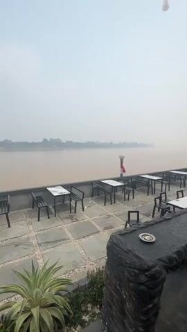 A still, overcast day with a hazy sky and calm, muddy water forms the backdrop to an empty outdoor seating area. Rows of tables and chairs, mostly black with white tops, are arranged on a tiled patio overlooking the water. A low stone wall separates the seating area from the river. In the foreground, a prickly green plant sits amidst some smaller foliage. String lights hang overhead, and a few lamps are visible along the water's edge. The scene is quiet and appears to be early morning or late afternoon due to the diffused light.