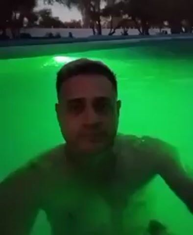 A man with short brown hair and a light complexion is shown from the chest up, swimming in a pool. He faces forward, looking directly at the camera, and moves slightly in the water. The pool is illuminated with green, then blue, lights, and the background reveals what appears to be an outdoor area at dusk, with trees and a fence visible.