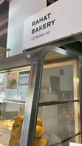 The video shows the inside of a bakery named "RAHAT BAKERY," which also has the Vietnamese translation for bakery, "Lò Bánh Mì." Displayed on the front of a stainless steel display case are loaves of bread. The camera pans right to reveal a man standing behind the counter. He is wearing glasses and has tattoos on his arms. In front of him are signs with pictures and prices of pastries in Vietnamese Dong. There are signs for chocolate rolls, lemon cheesecake, cottage cheese, and another dessert. Behind the man, there is a kitchen with stainless steel counters, cabinets, and a ventilation hood. A small red lantern hangs from the ceiling. The man in the video appears to be preparing to take an order.