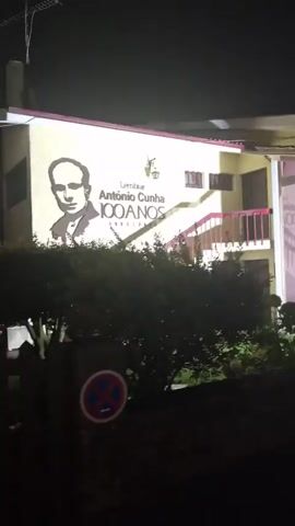 The video shows a building at night with a mural of a man's face on the left side, along with text that appears to say "Lembrar António Cunha 100 ANOS 1922 2022". To the right, there is a glass-enclosed display with several white statues of people in historical clothing. A dark-colored car is parked in front of the display, partially obscuring the view of the statues and the building. The environment appears to be outdoors with some vegetation in the foreground. The lighting suggests artificial illumination from the building and possibly streetlights.