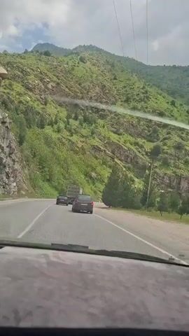 The video shows a car driving on a paved road with a mountain on the left. The mountain is green with trees and some exposed rock. There are other vehicles on the road, including a car and a truck. The sky is cloudy. The video appears to be shot from inside a car. Music is playing in the background.