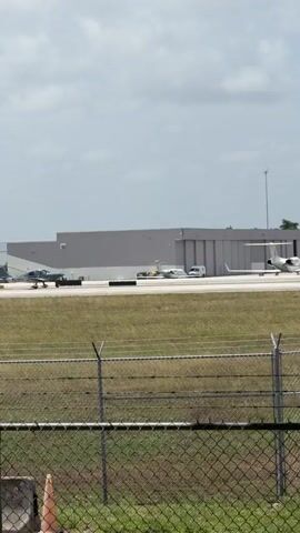 A fixed-wing aircraft taxis on a runway. The aircraft appears to be a small, twin-engine propeller plane. In the background, a larger, white jet aircraft is parked on the tarmac. Other aircraft and airport buildings are visible in the distance. The scene is set against a partly cloudy sky. A chain-link fence with barbed wire runs along the foreground. The video is shot from a stationary perspective.