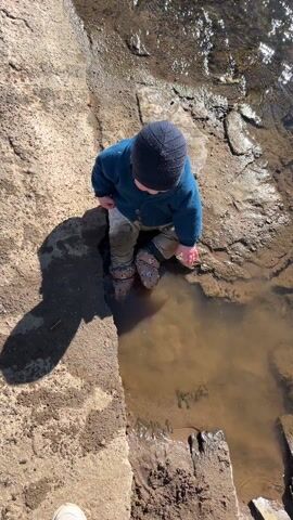 A toddler in a blue sweater and a knit hat stands near a puddle of muddy water. The child reaches into the water with one hand. The child is wearing sandals and standing on a stone surface. The puddle is surrounded by dirt and rocks. The sun is shining and there are reflections on the water.