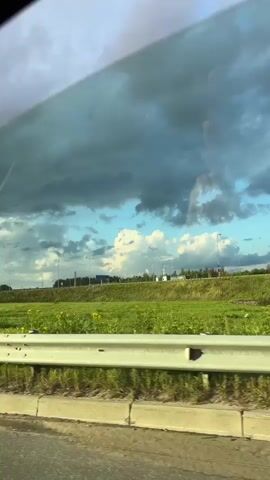 The video shows a view from inside a vehicle looking out a window. The window is tinted, giving the sky a slightly darker hue. Outside, there is a grassy field with some wildflowers. A metal guardrail runs along the edge of the field. In the distance, there is a highway with some traffic, including cars and possibly trucks. There are also some tall poles with lights along the highway. The sky is filled with dramatic, dark clouds, but there are also patches of blue sky and white clouds. The overall impression is of a landscape viewed during a transitional period, possibly before or after a storm.