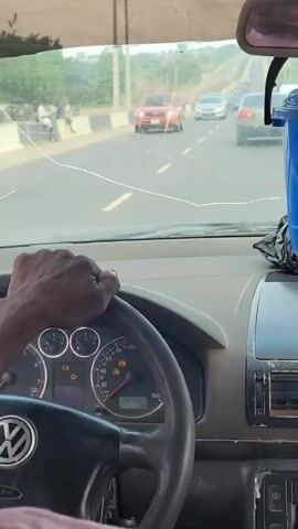 The video provides a first-person perspective from inside a moving car, likely a Volkswagen due to the logo on the steering wheel, on a road in Uke, Nigeria. The driver's dark-skinned hand is visible on the steering wheel.

The dashboard, which shows signs of wear and tear, is in the foreground, with various warning lights illuminated on the instrument cluster, including what appears to be a check engine light, battery light, and oil pressure light. The odometer reads 20430.

A prominent blue plastic bucket, possibly containing items like sachets or small bottles, is placed on the dashboard and hangs from the rearview mirror. Some text or labels are visible on the bucket through a transparent covering, but they are not clearly legible. The rearview mirror is also visible.

Through the cracked windshield, a multi-lane road is seen ahead with moderate traffic. Several cars, including a red pickup truck and various sedans, are visible moving in the same direction. The surrounding landscape appears to be somewhat rural or suburban with distant trees or hills. The vehicle is driving during the daytime under bright conditions.