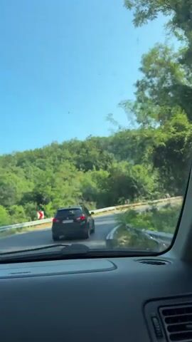 The video shows the view from inside a car, looking out through the windshield at a road curving through a forest. A dark-colored SUV is driving ahead on the road. The sky is blue and clear, and the sun is shining, casting light on the trees. There are curve warning signs on the side of the road. The interior of the car is visible in the foreground, with the dashboard and air vents. The overall impression is a scenic drive on a sunny day.