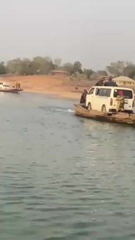 The video shows two small boats, each carrying a white van and several people, traveling across a river. The boats are simple wooden rafts, and the vans appear to be tightly secured on them. People are standing on the rafts alongside the vans, some dressed in local attire. The riverbank is visible, featuring a sandy shore, some trees, and a few buildings in the distance. The sky is overcast, creating a somewhat hazy atmosphere. The boats are moving towards the opposite shore where people are also standing.