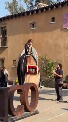 In a medium shot outdoors, a woman stands on stilts dressed in a floor-length, black and gold gown with a man playing a trumpet. The woman is on the left and dressed in period clothing, while the man is standing on the right with a music stand in front of him. They are both facing the camera, slightly turned to each other, with the woman holding a microphone. A large, rustic metal sign with the letters "CO" is in the foreground. The backdrop features a two-story building of tan stucco with arched windows and decorative shutters. There are a few other people walking by in the background. The lighting is bright and sunny.