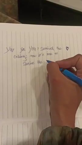 A close-up shot shows a person's hand holding a blue pen and writing on a lined notebook. The handwriting is in cursive and reads "You yes You? Survives the ending; now it is time to Survive the healing." A small heart symbol is drawn next to the word "ending." The notebook is open on a surface that appears to be a table or counter. The lighting suggests an indoor setting, possibly a home.