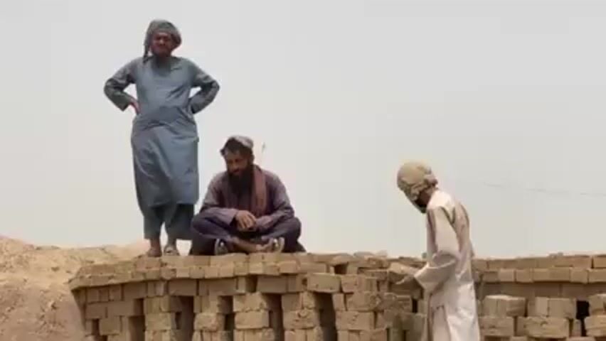 Three men are outside, in what appears to be a hot, dusty environment. Two of the men have head coverings. One man stands with his hands on his hips, observing the scene. Another man sits cross-legged atop a pile of bricks. The third man, covered in dust, is actively stacking bricks.