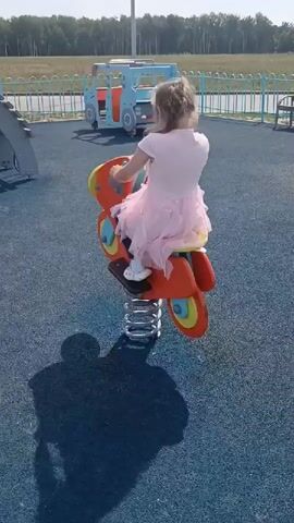 A young girl in a pink dress is playing on a spring-loaded toy motorcycle on a playground. The playground has a blue rubberized surface and a fence in the background. In the background, there are trees, a white truck, and another blue playground vehicle. The girl is bouncing on the toy and appears to be enjoying herself. The sun is shining, casting shadows on the ground.